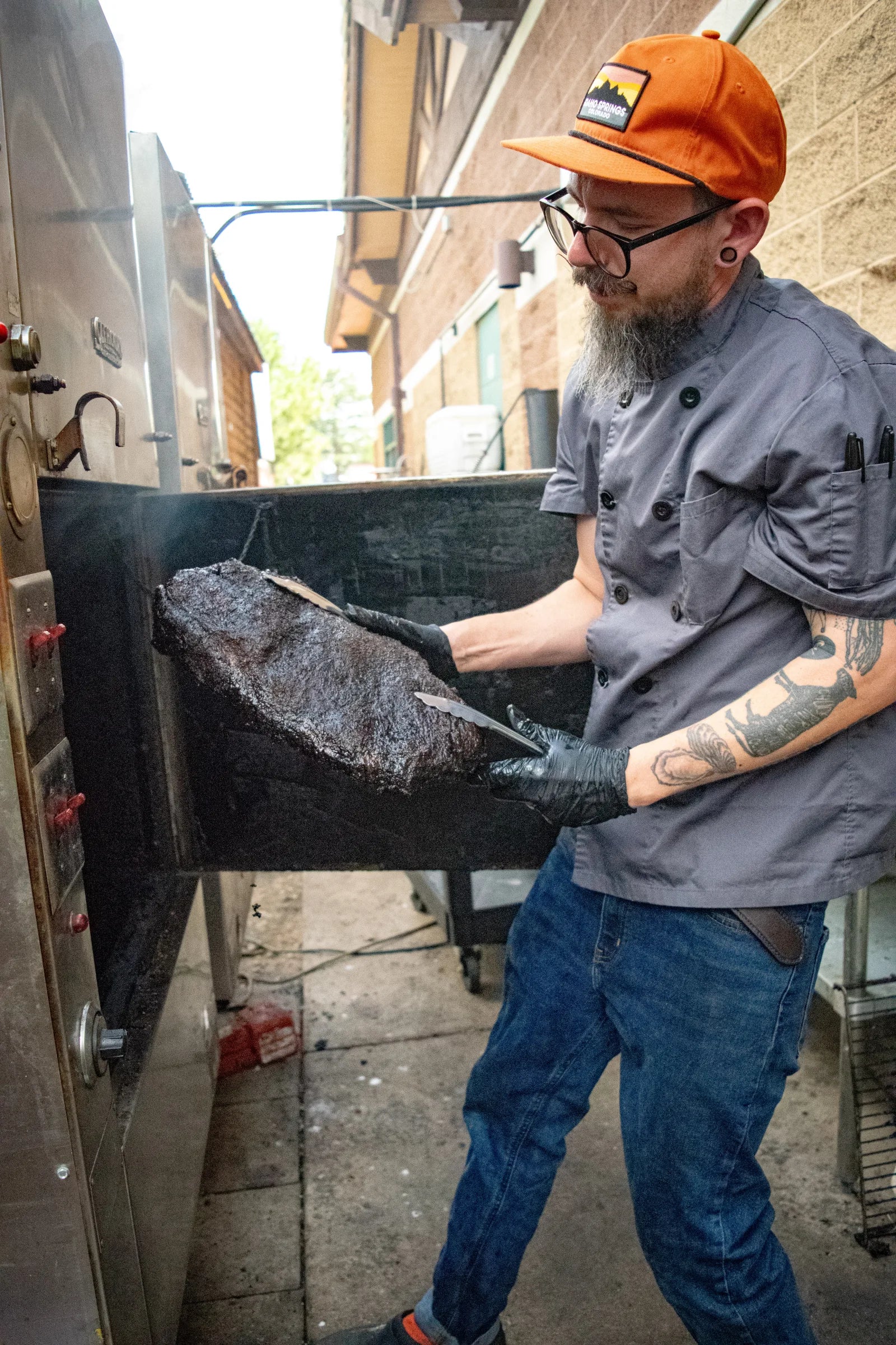 Bearded cook wearing glasses and an orange cap, pulling a large dark smoked brisket out of a commercial smoker on a loading dock, with smoke in the air.