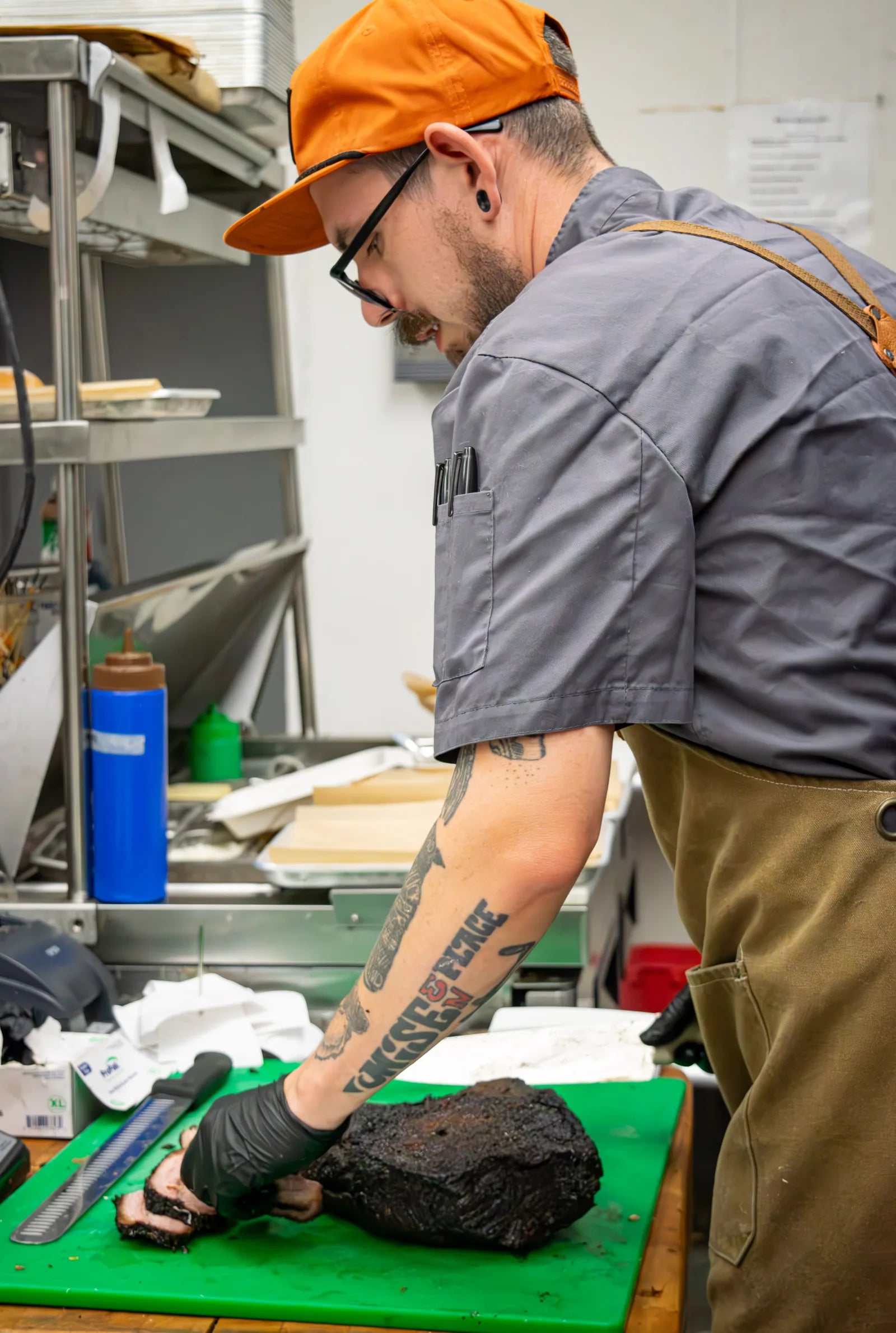 Cook wearing an orange cap and glasses slicing a dark bark-crusted smoked brisket on a green cutting board.