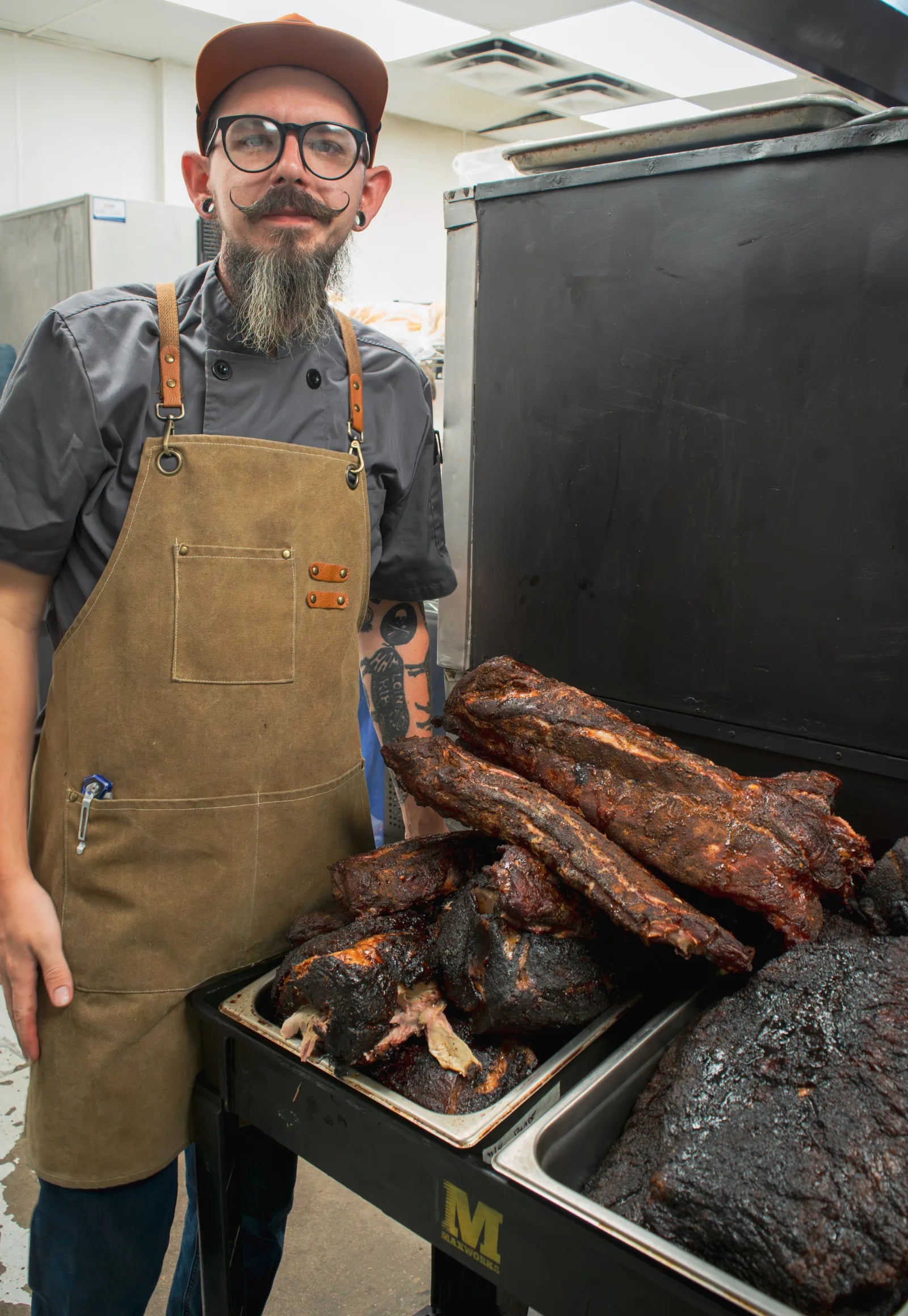 Smiling cook with a long gray beard, round glasses, and a tan leather apron standing beside a black smoker loaded with trays of smoked rib racks and brisket.