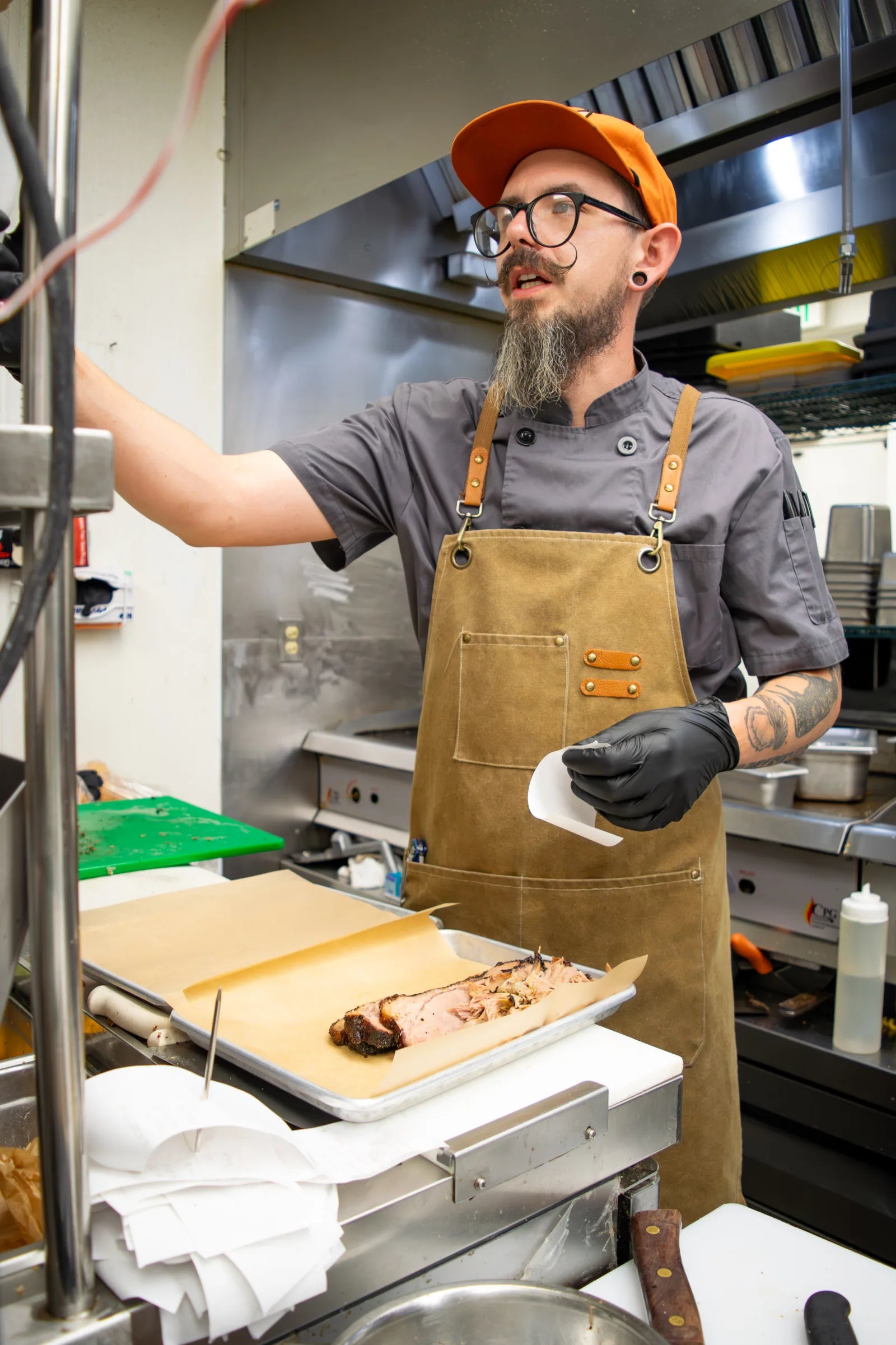 Bearded cook wearing round glasses, an orange cap, and a tan leather apron, standing at a kitchen station with a tray of sliced and pulled brisket.
