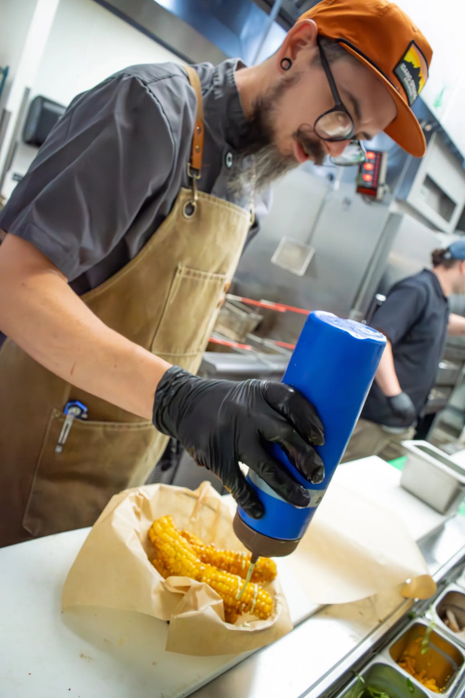 Bearded cook wearing glasses, an orange cap, and a tan leather apron squeezing sauce from a blue bottle onto grilled corn.
