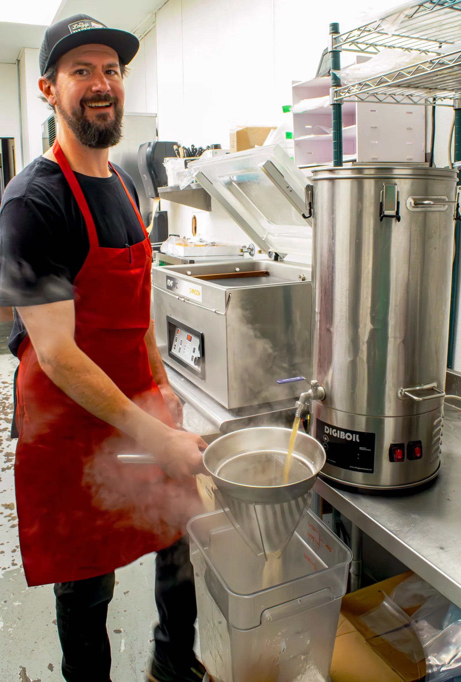 Smiling bearded cook wearing a black Lazy Butcher cap and red apron, pouring steaming liquid through a strainer into a container.