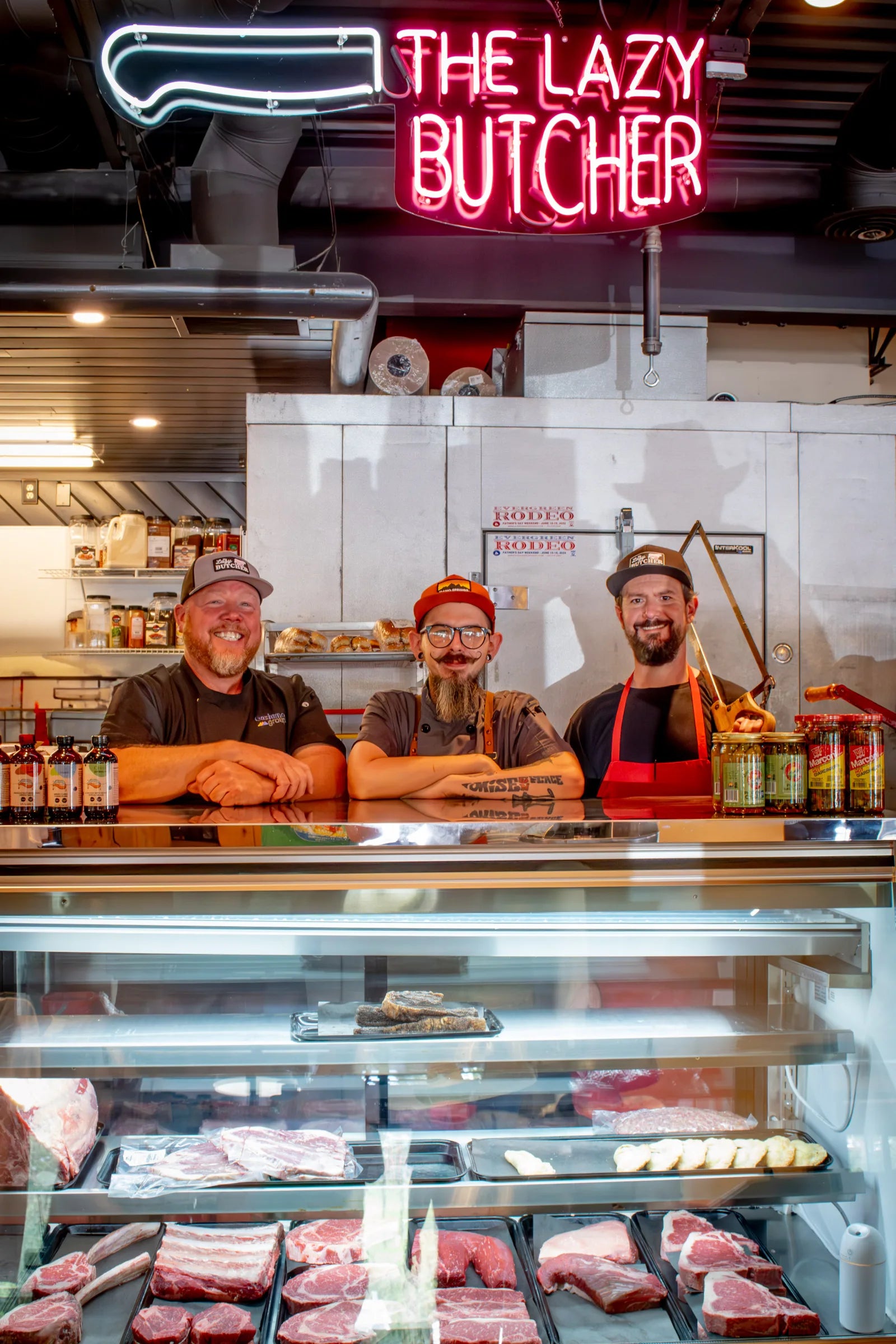 Three smiling men in Lazy Butcher caps and aprons leaning on the counter behind the glass meat case, with the glowing neon 'The Lazy Butcher' knife sign mounted above them.