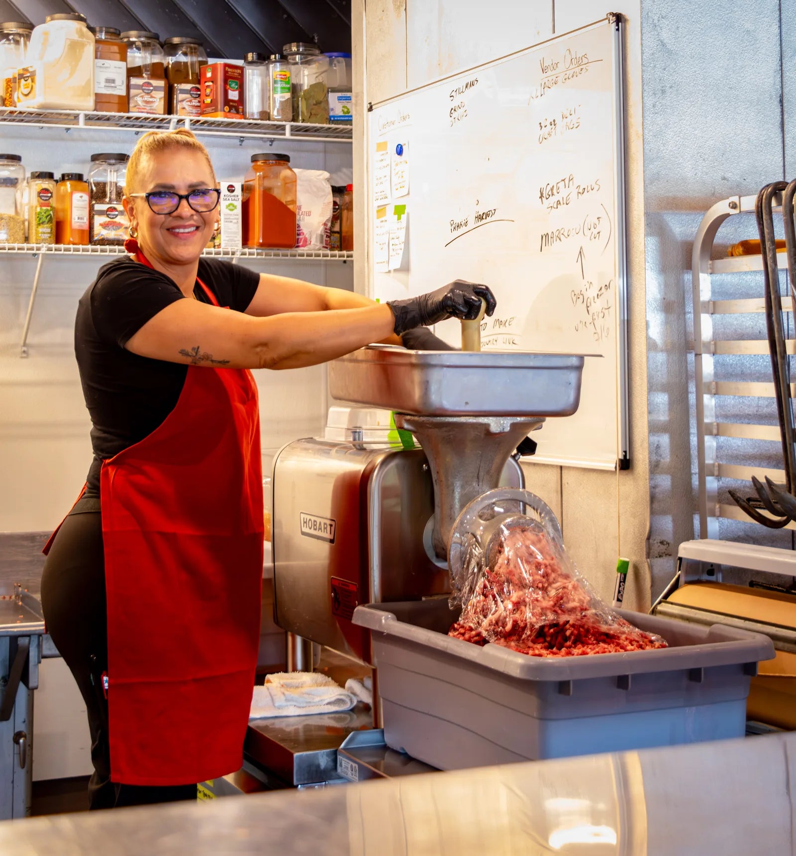 Smiling woman with glasses and a red apron operating a Hobart meat grinder, with ground meat filling a bin below.
