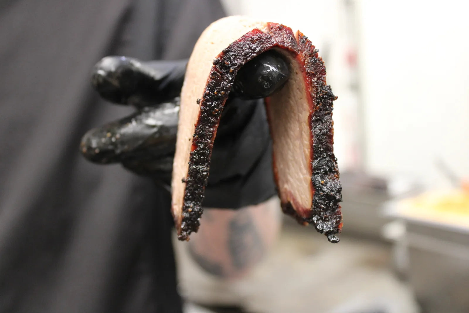 A gloved hand holds up a folded slice of smoked brisket, showing a deep pink smoke ring and dark bark crust.