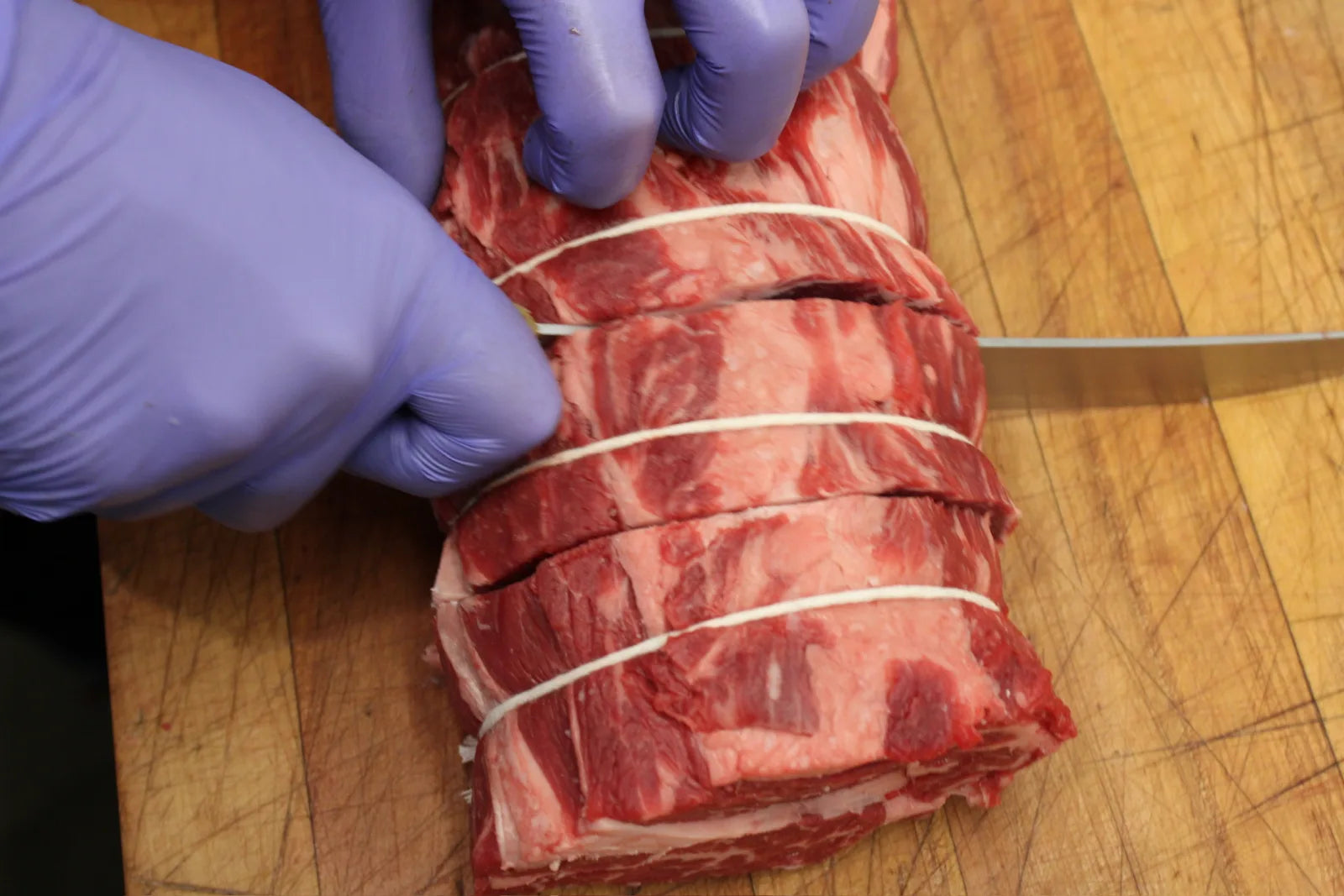 A butcher's gloved hands guide a knife through a beef roast tied with butcher's twine, slicing it into individual steaks.