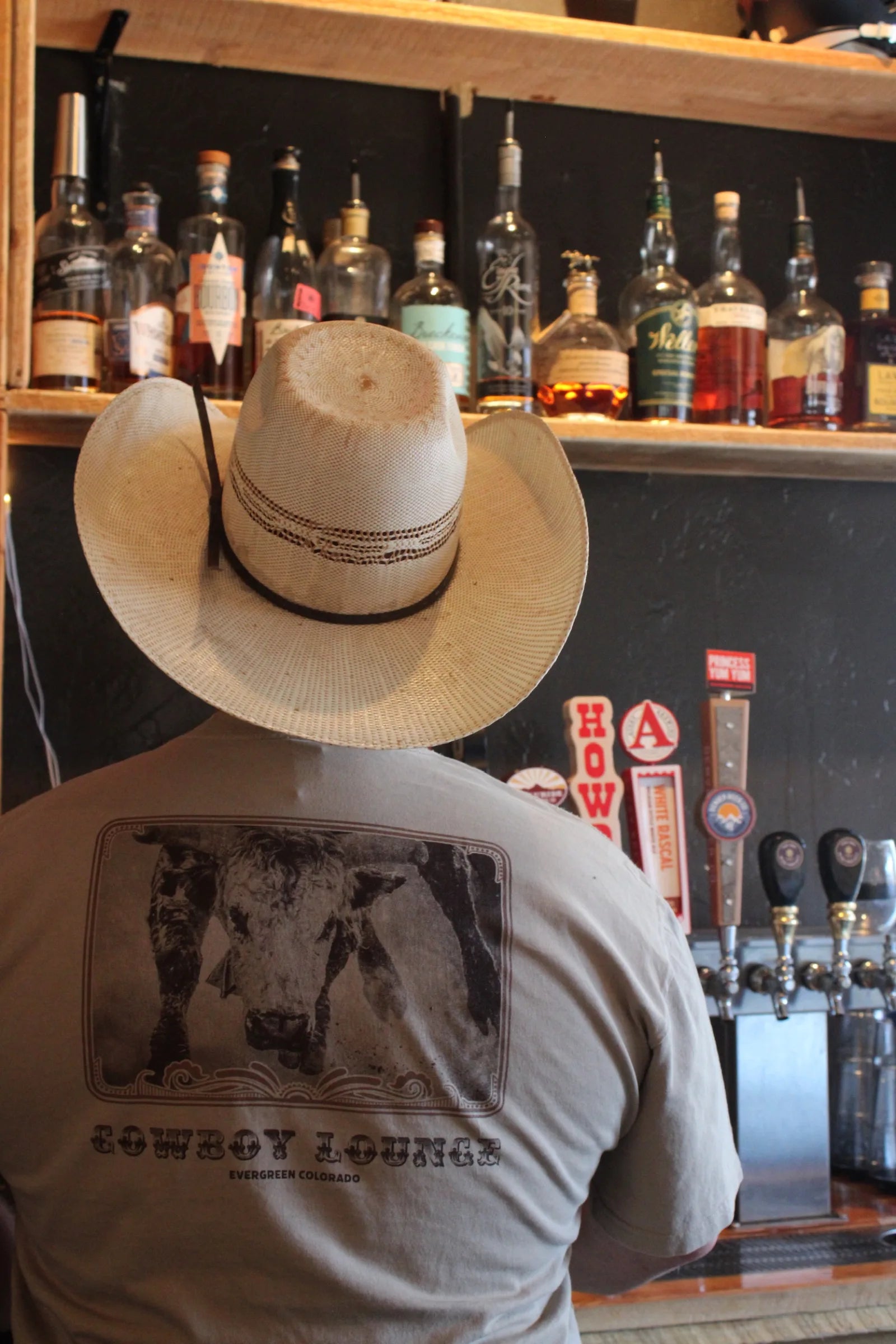 A man in a straw cowboy hat and Cowboy Lounge t-shirt with a cattle graphic stands facing the bar's shelves of whiskey bottles and beer taps.