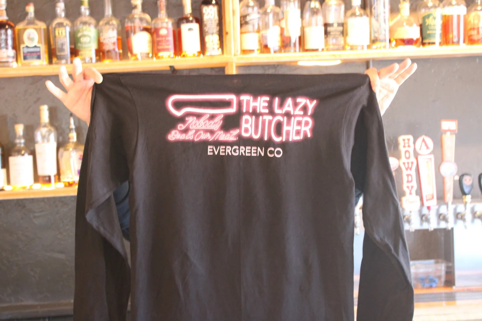 Hands hold up a black t-shirt with The Lazy Butcher's neon-style knife logo in front of a bar shelf of whiskey bottles.