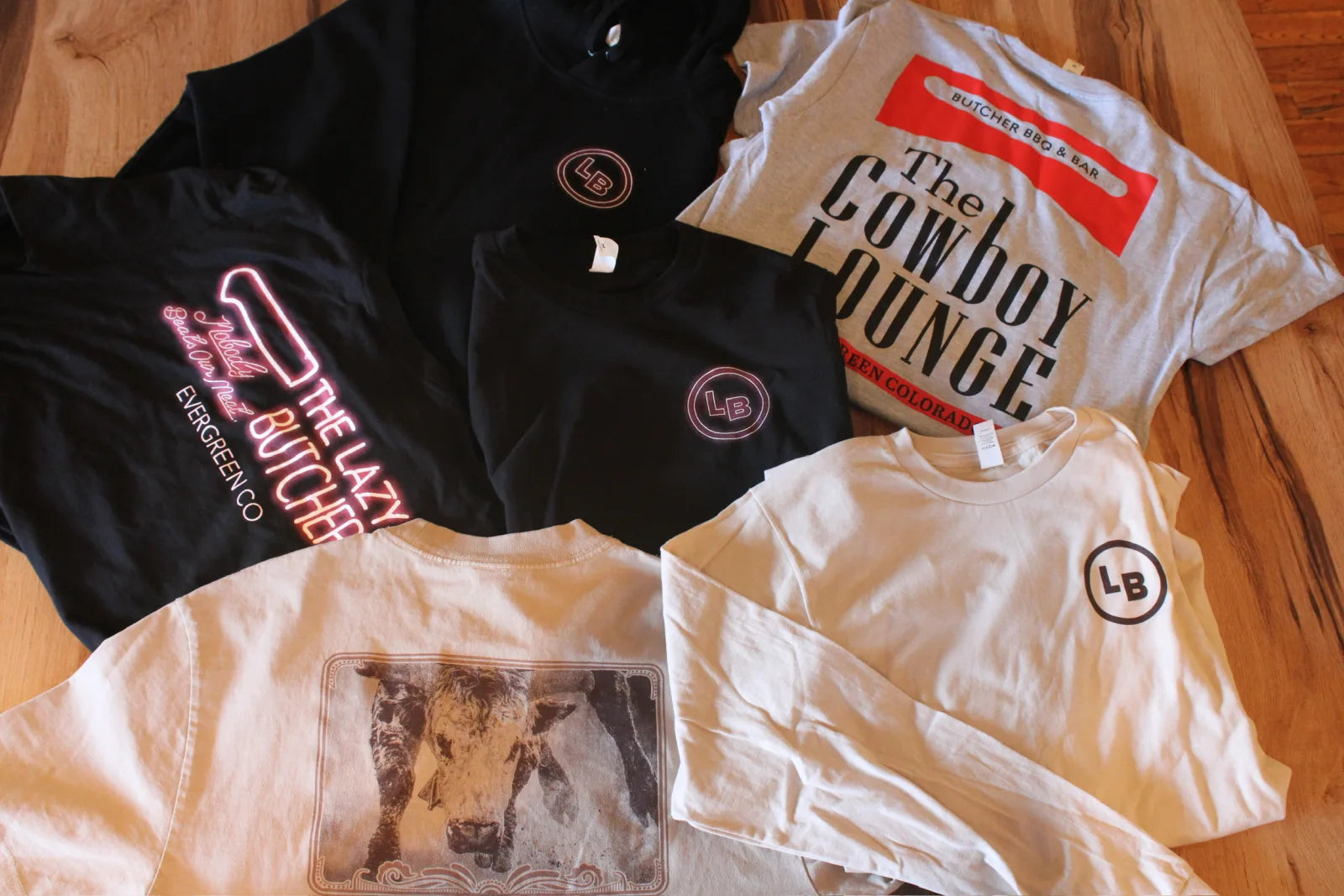 A spread of branded t-shirts and a hoodie for The Lazy Butcher and The Cowboy Lounge laid out on a wood floor.