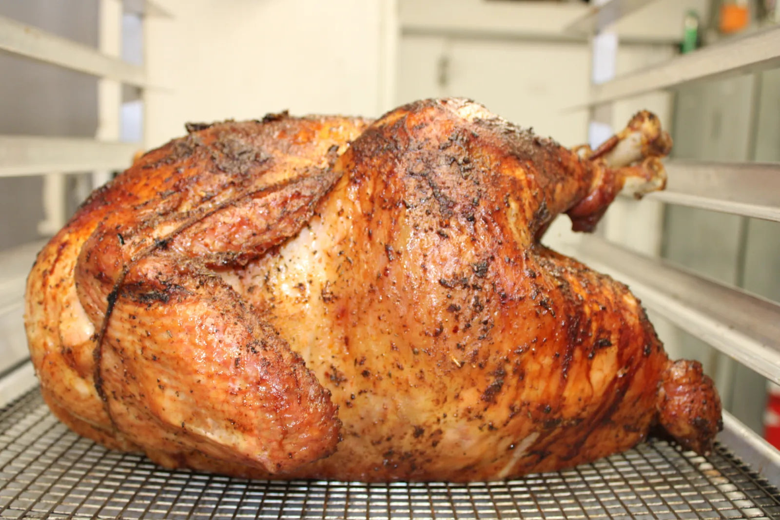 A whole smoked turkey with a deep mahogany, spice-crusted skin resting on a wire rack in a walk-in cooler.