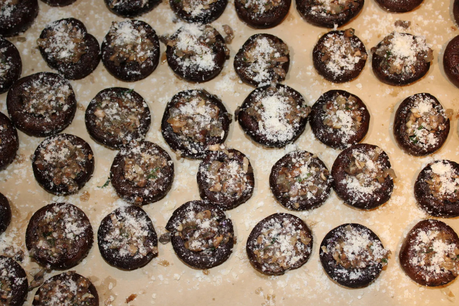 Rows of mushroom caps filled with a savory stuffing and topped with grated parmesan, arranged on a sheet pan.