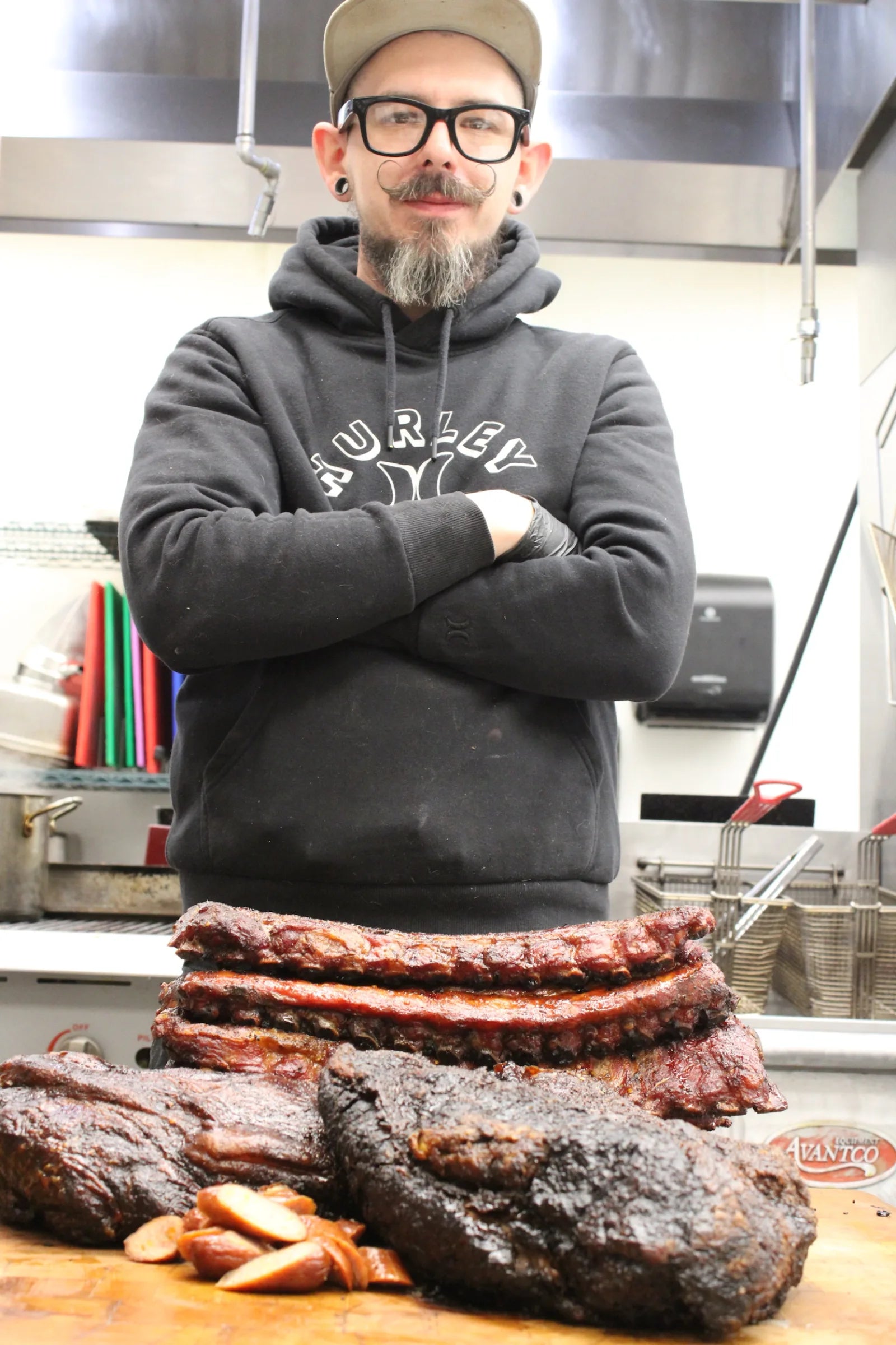 A bearded kitchen staff member in glasses and a backwards cap stands with arms crossed behind a spread of smoked brisket, ribs, and sliced sausage.