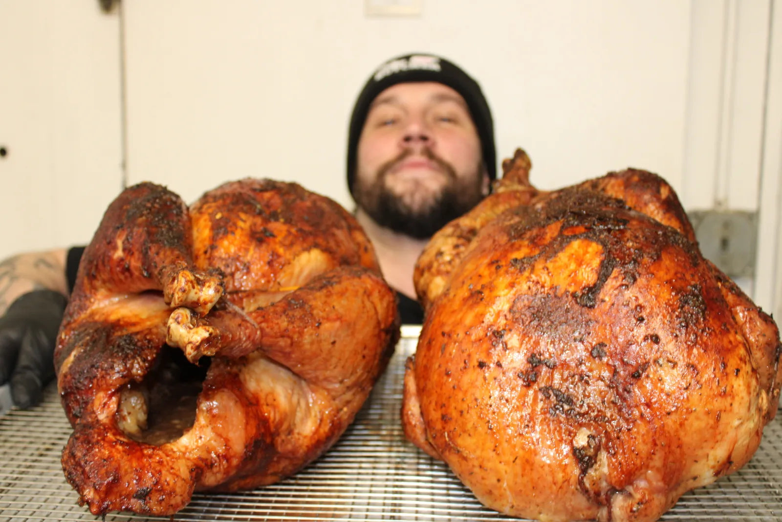 A bearded staff member in a beanie poses behind two whole smoked turkeys with dark, spice-rubbed skin on a cooling rack.