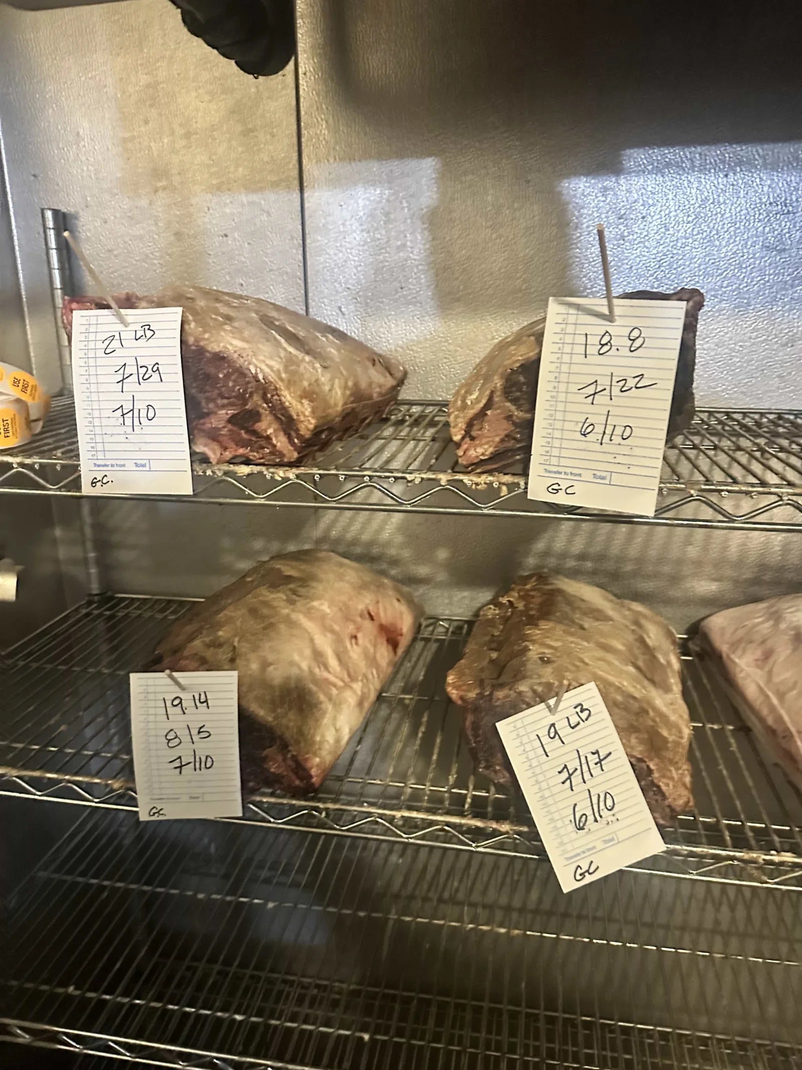 Beef primals dry-aging on cooler shelves, each with a hand-written tag noting weight and date.