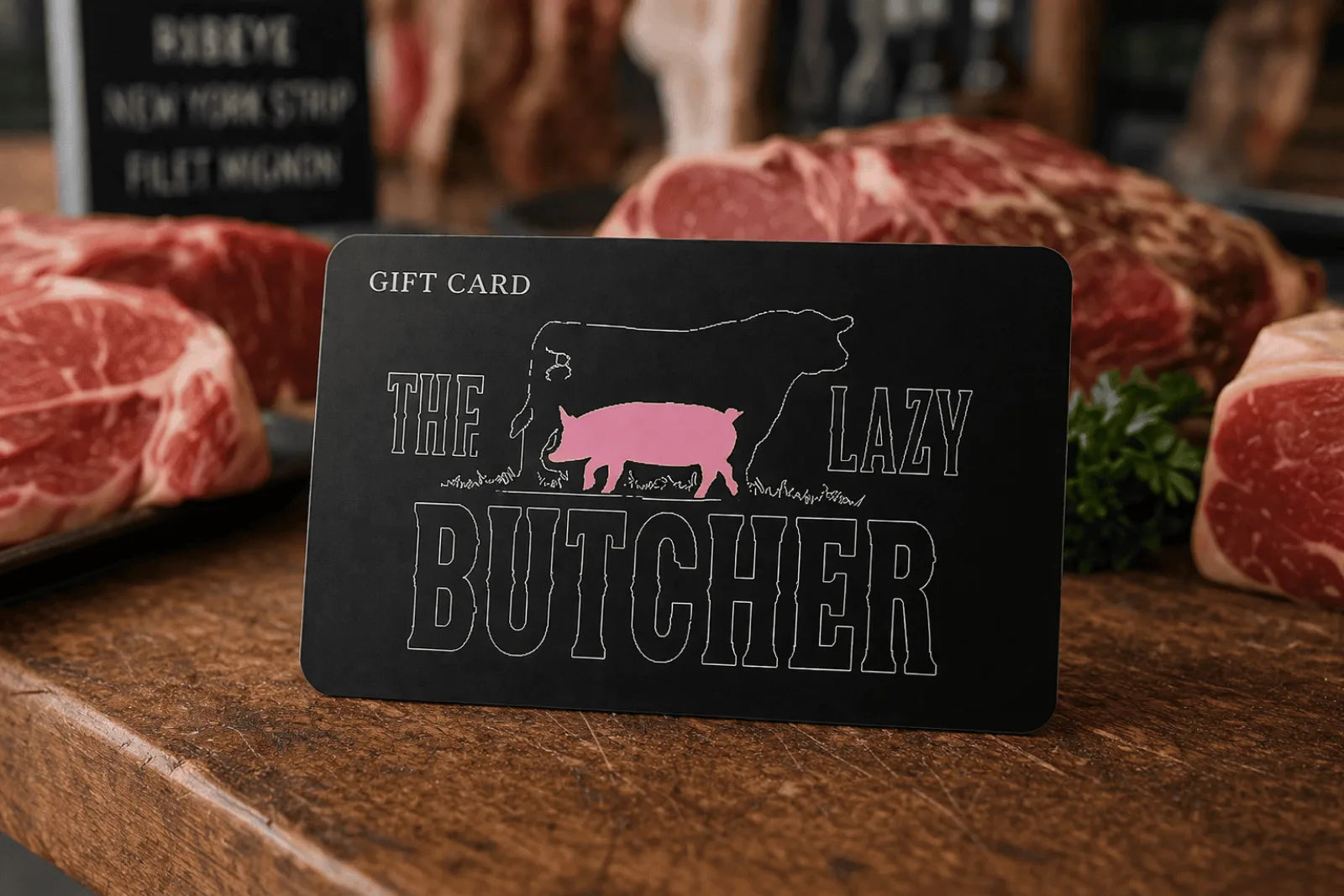 A Lazy Butcher gift card standing on a wood counter with dry-aged steaks in the butcher case behind it.