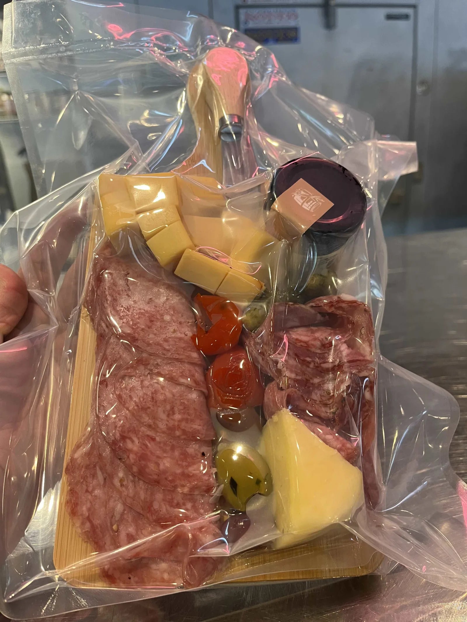 A vacuum-packed charcuterie board with sliced salami, cubed cheese, olives, and a jar of preserves.