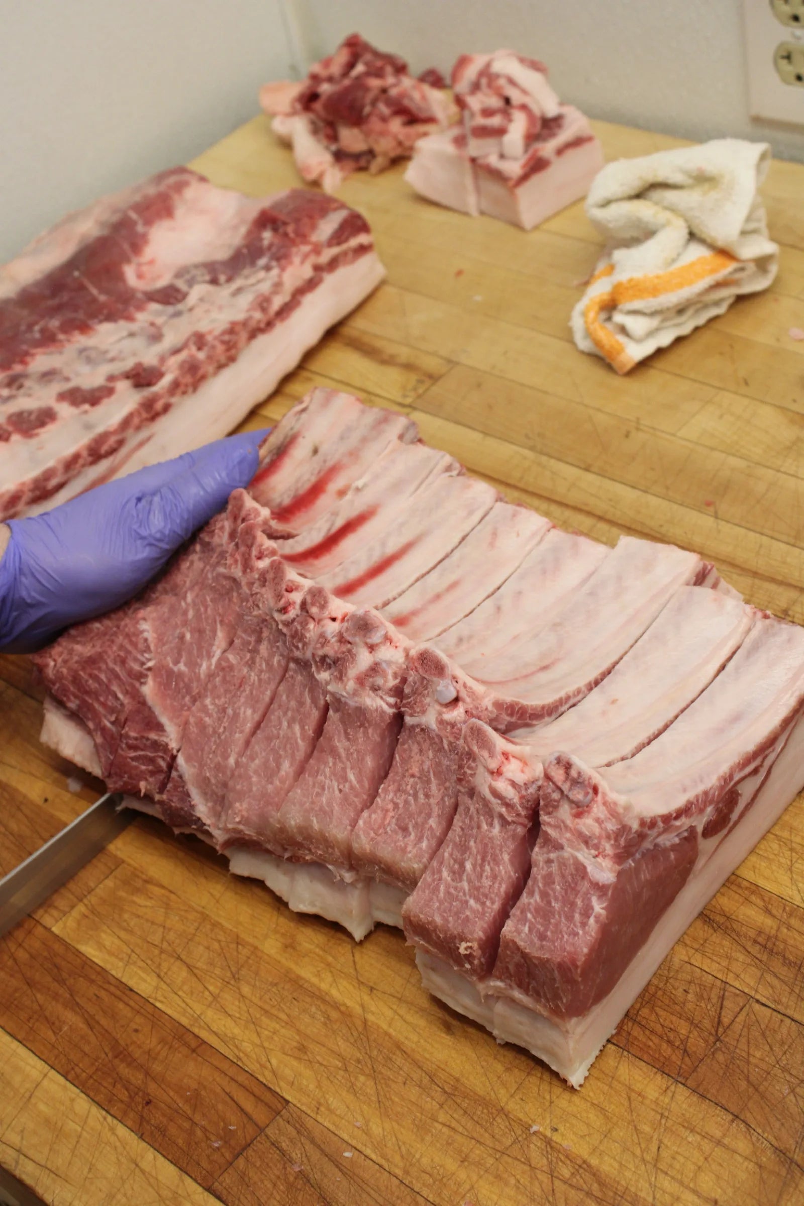 Raw pork belly and a trimmed rib rack on a wooden butcher block, with a gloved hand at the edge of frame.