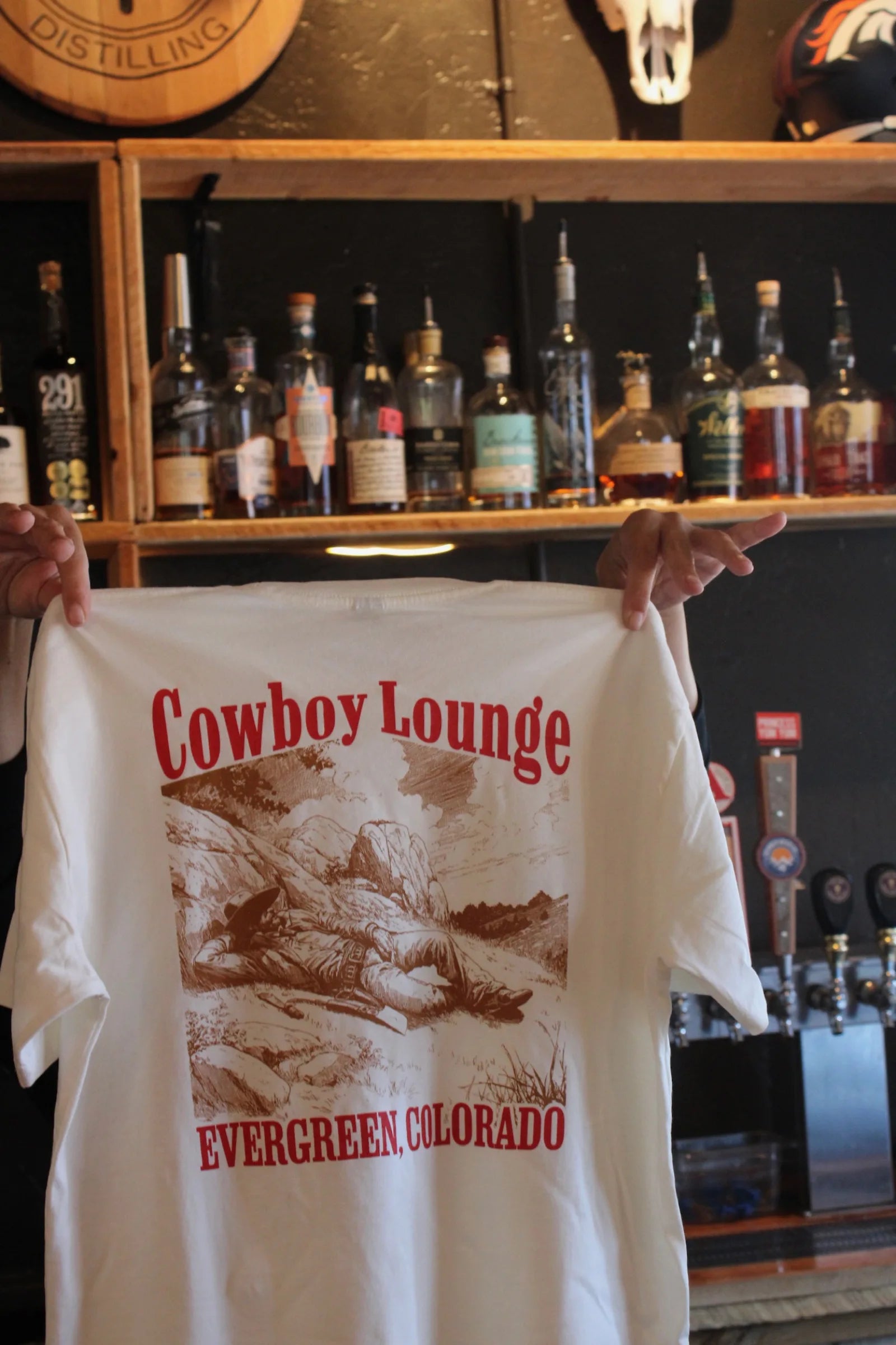 A white t-shirt reading 'The Cowboy Lounge, Evergreen Colorado' held up in front of the back bar shelves.