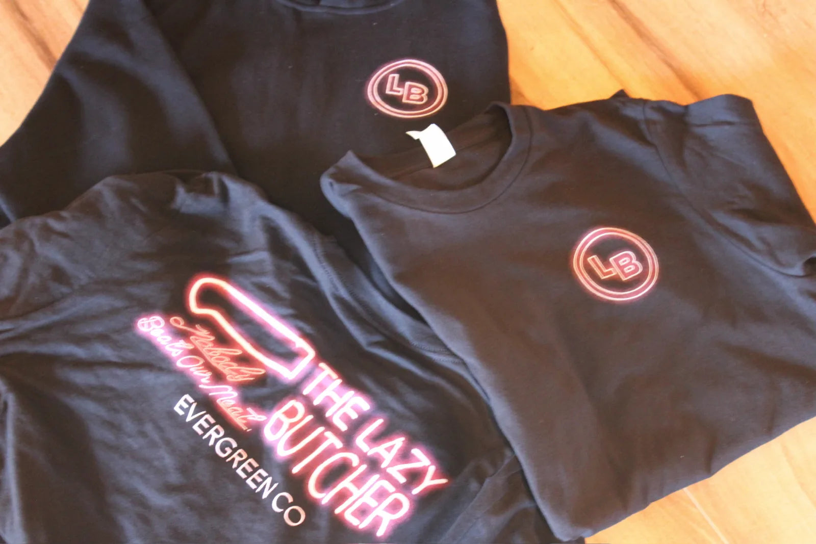 Folded black Lazy Butcher branded t-shirts on a wood table.
