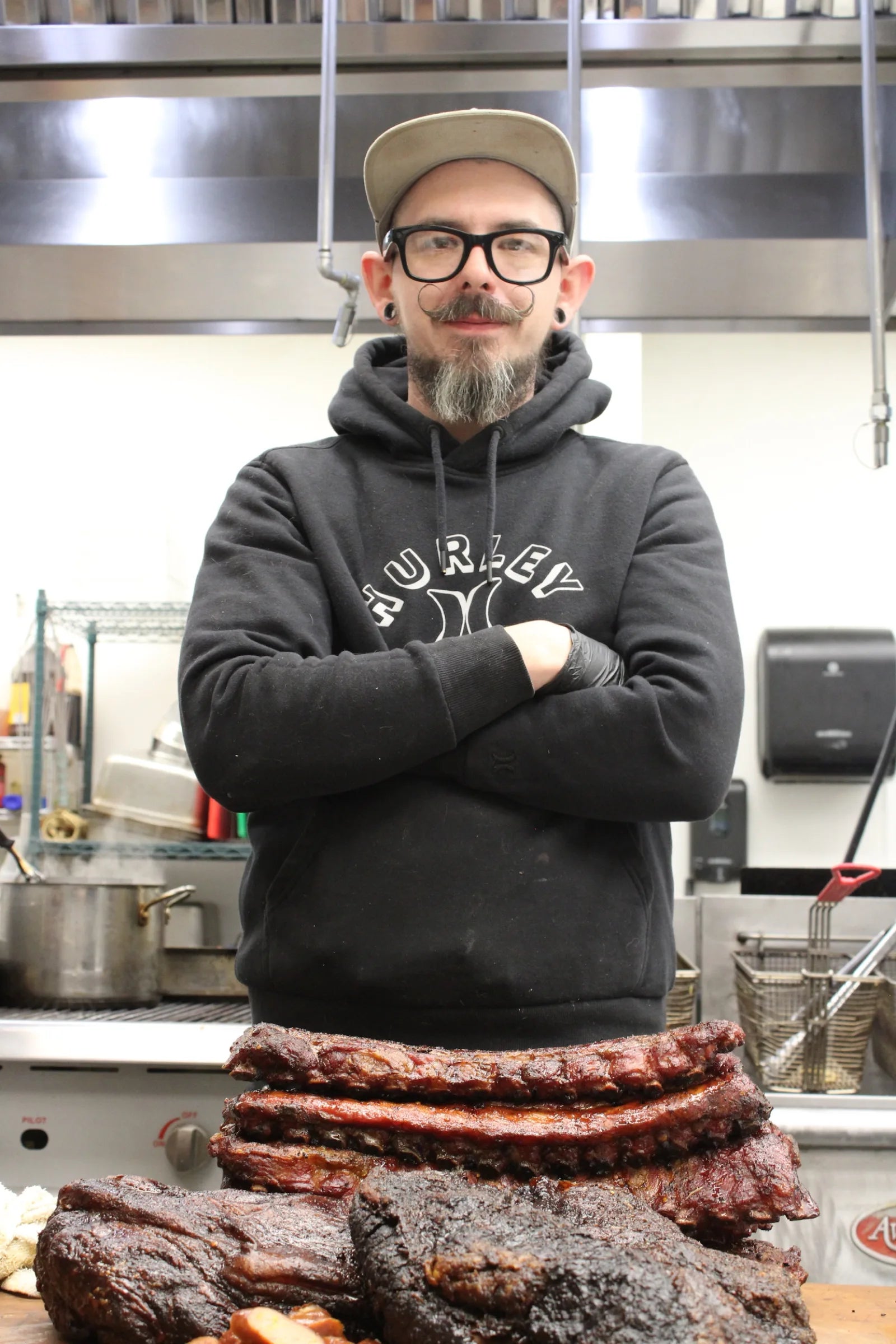 A bearded cook in glasses and a gray hoodie stands with arms crossed behind a full spread of smoked ribs and brisket.