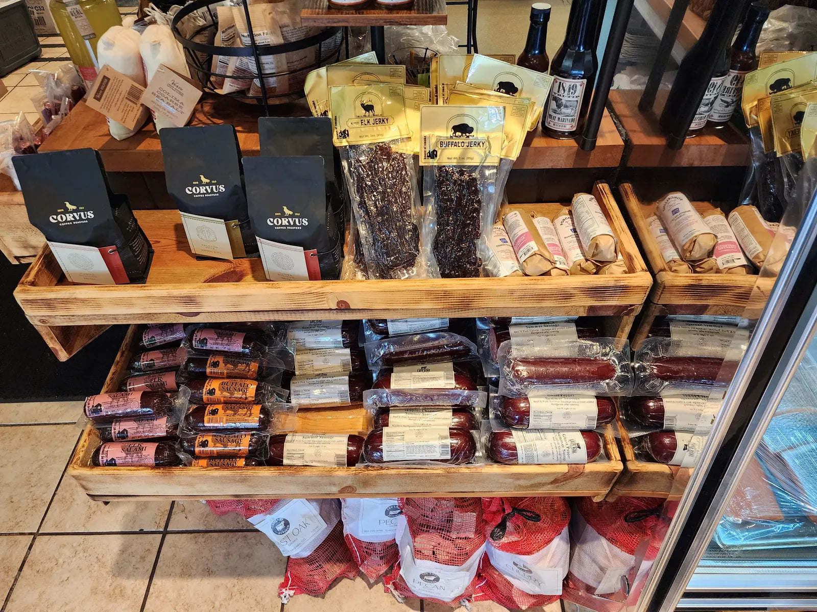 Elk and buffalo jerky, elk salami, summer sausages, and Corvus coffee on wooden crates, with bags of smoking wood below