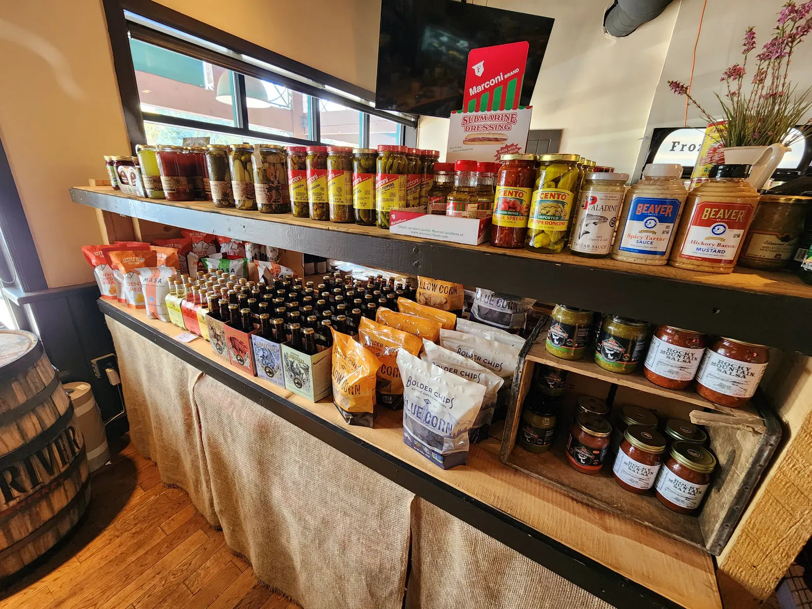 A shelf of pickles, giardiniera, peppers, mustards, Rocky Mountain Salsa, Bolder Chips, and bottled craft soda